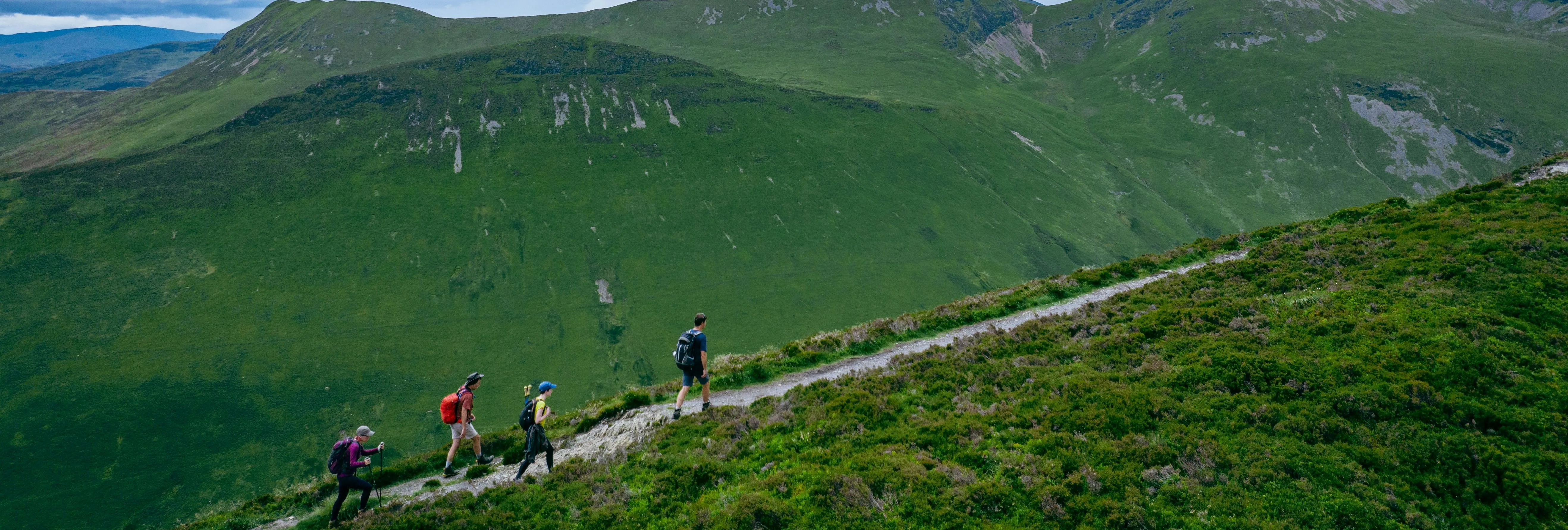 Four hikers walking along a narrow trail across a green mountain hillside beneath a cloudy sky.