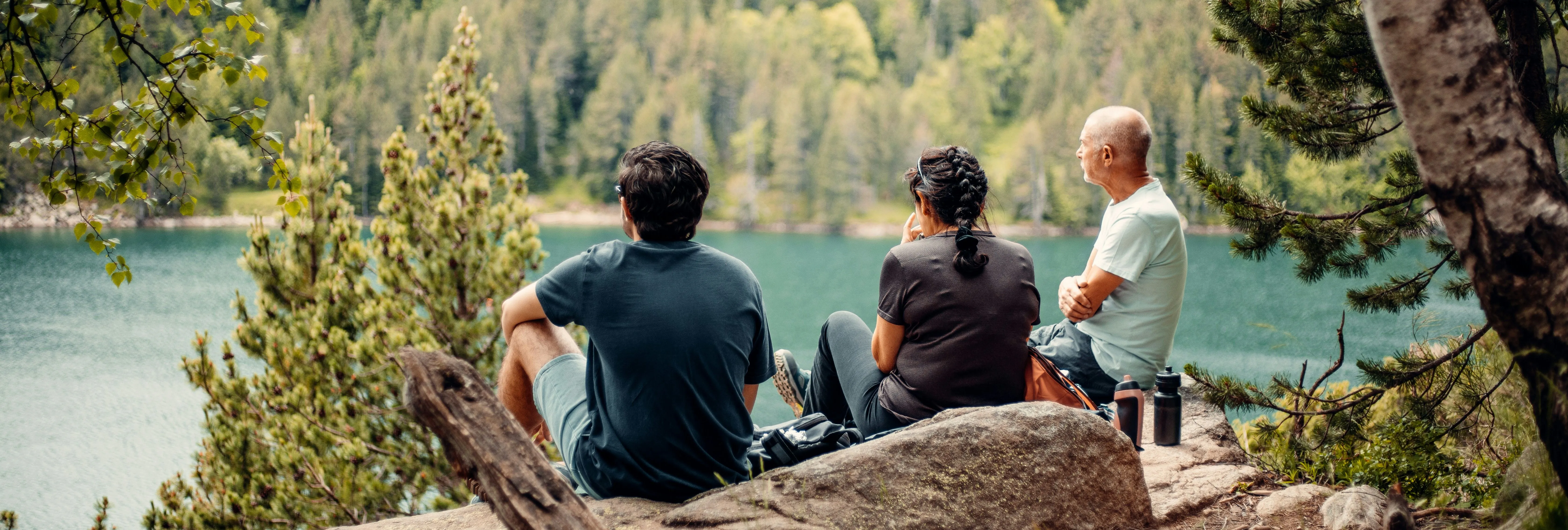 Three hikers resting on rocks beside a turquoise mountain lake surrounded by dense forest.