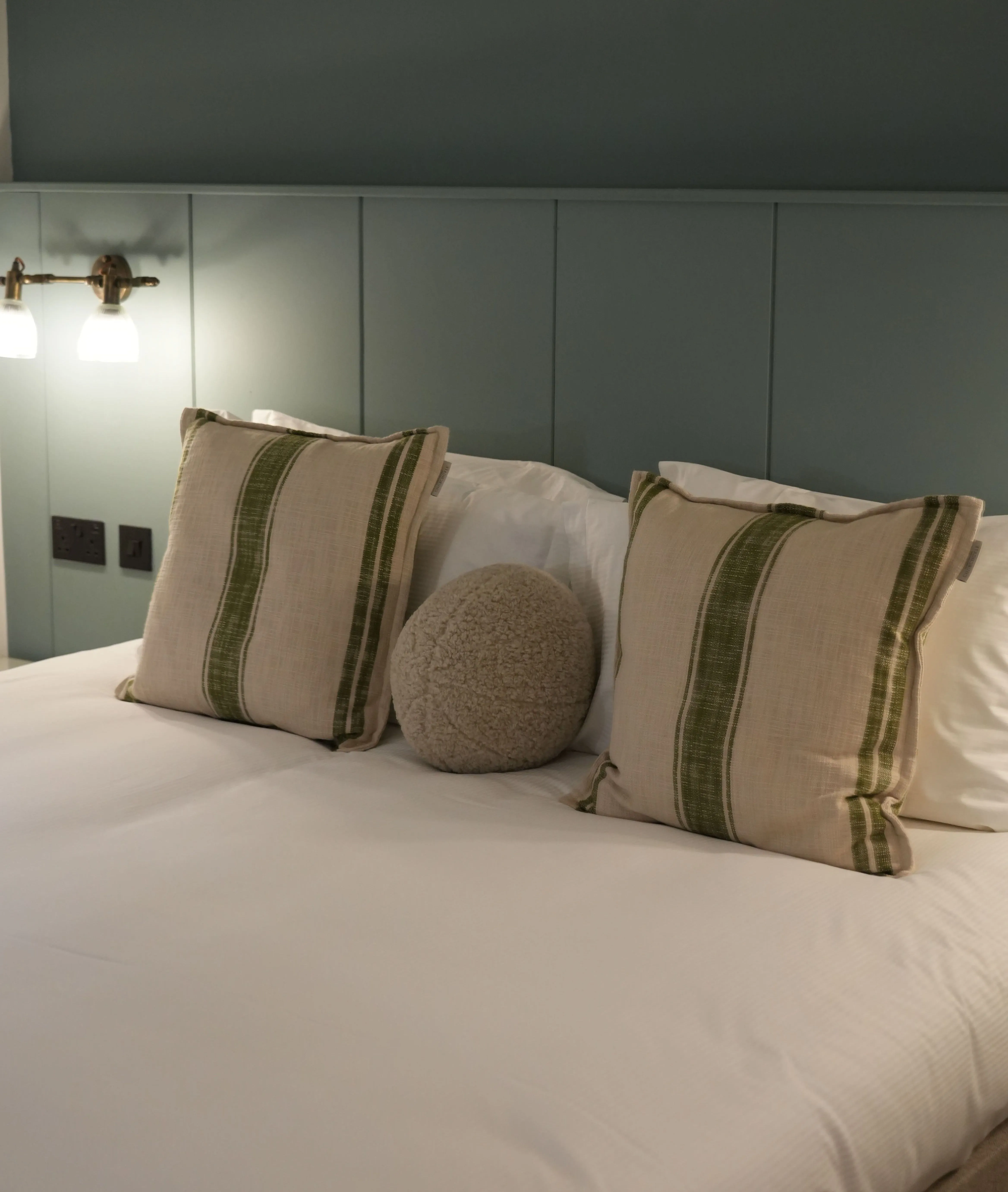 Cosy bedroom with a neatly made white bed, striped cushions and a green panelled headboard.