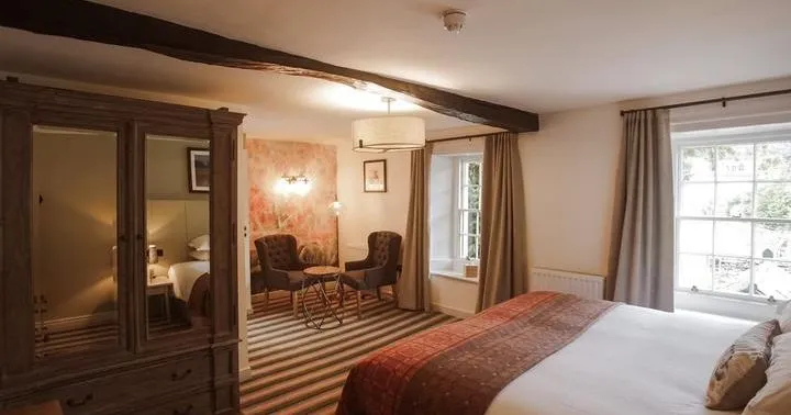 Traditional hotel bedroom with a large bed, dark wooden wardrobe, exposed ceiling beams and a small seating area.