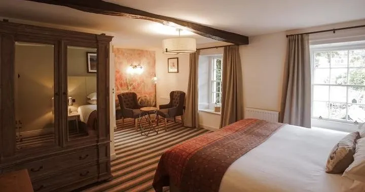 Traditional hotel bedroom with a large bed, dark wooden wardrobe, exposed ceiling beams and a small seating area.
