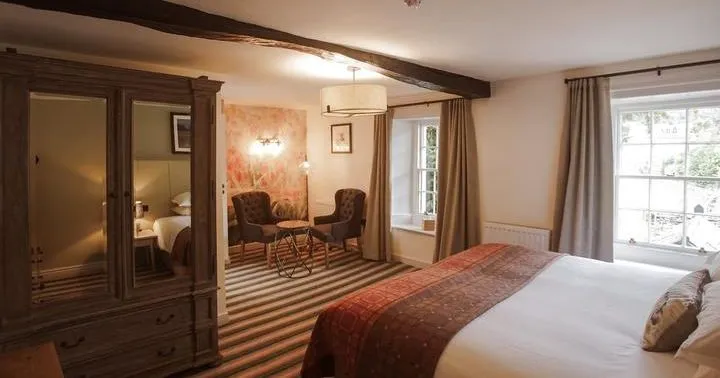 Traditional hotel bedroom with a large bed, dark wooden wardrobe, exposed ceiling beams and a small seating area.
