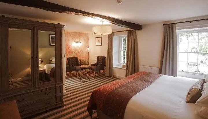 Traditional hotel bedroom with a large bed, dark wooden wardrobe, exposed ceiling beams and a small seating area.