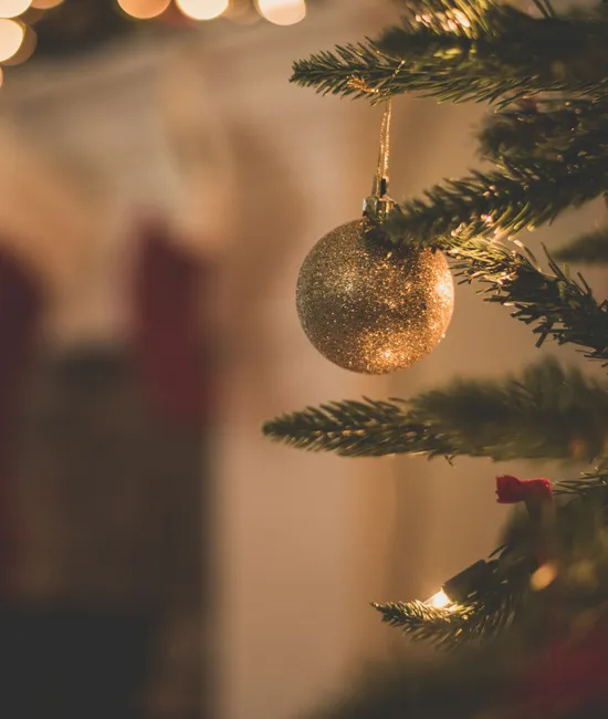 Gold bauble hanging from a decorated Christmas tree with warm blurred lights and wrapped gifts in the background.