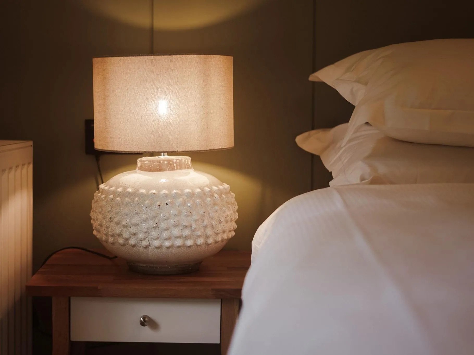 Warmly lit bedside table with a textured ceramic lamp beside a neatly made bed