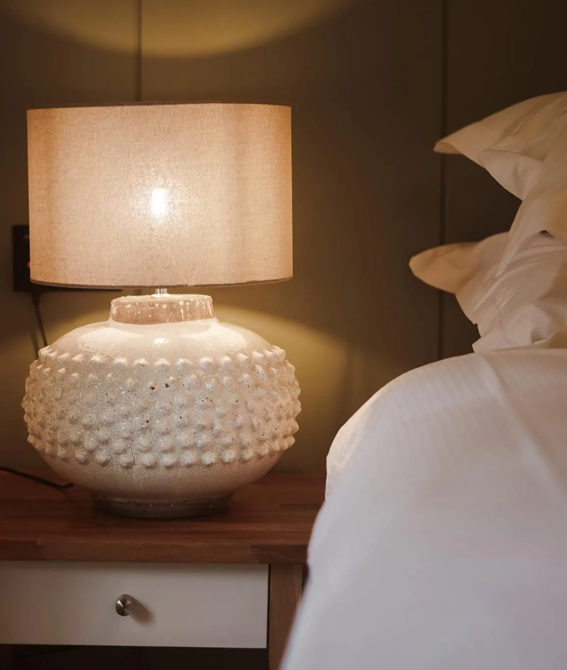 Warmly lit bedside table with a textured ceramic lamp beside a neatly made bed