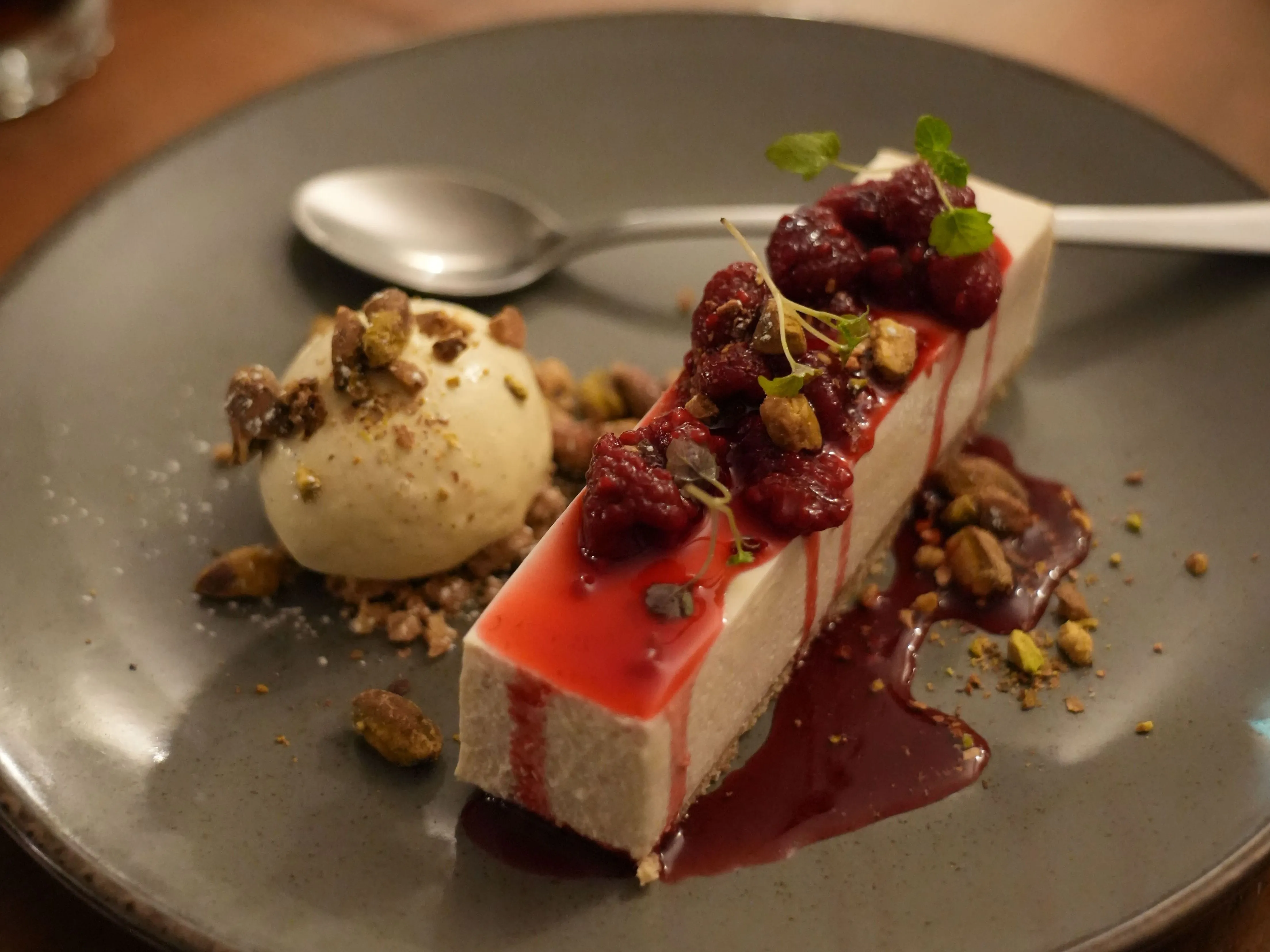 Slice of creamy dessert topped with berry compote, pistachios and a scoop of ice cream on a grey plate.