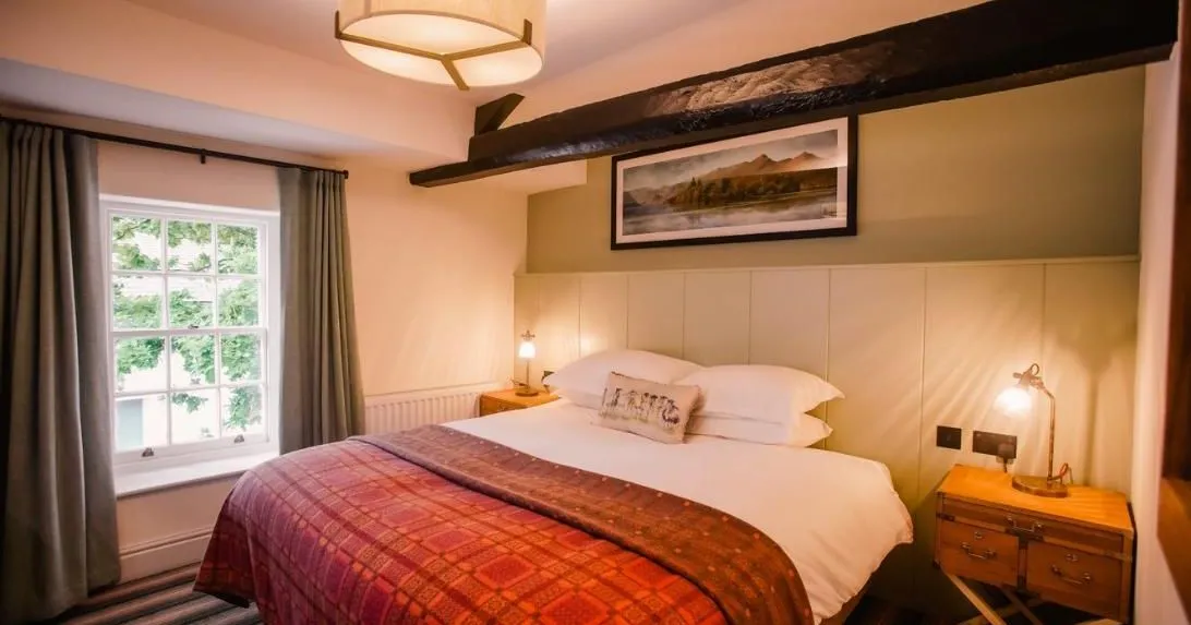 Cosy bedroom with a double bed, red patterned throw, exposed dark timber beam and warm bedside lighting.