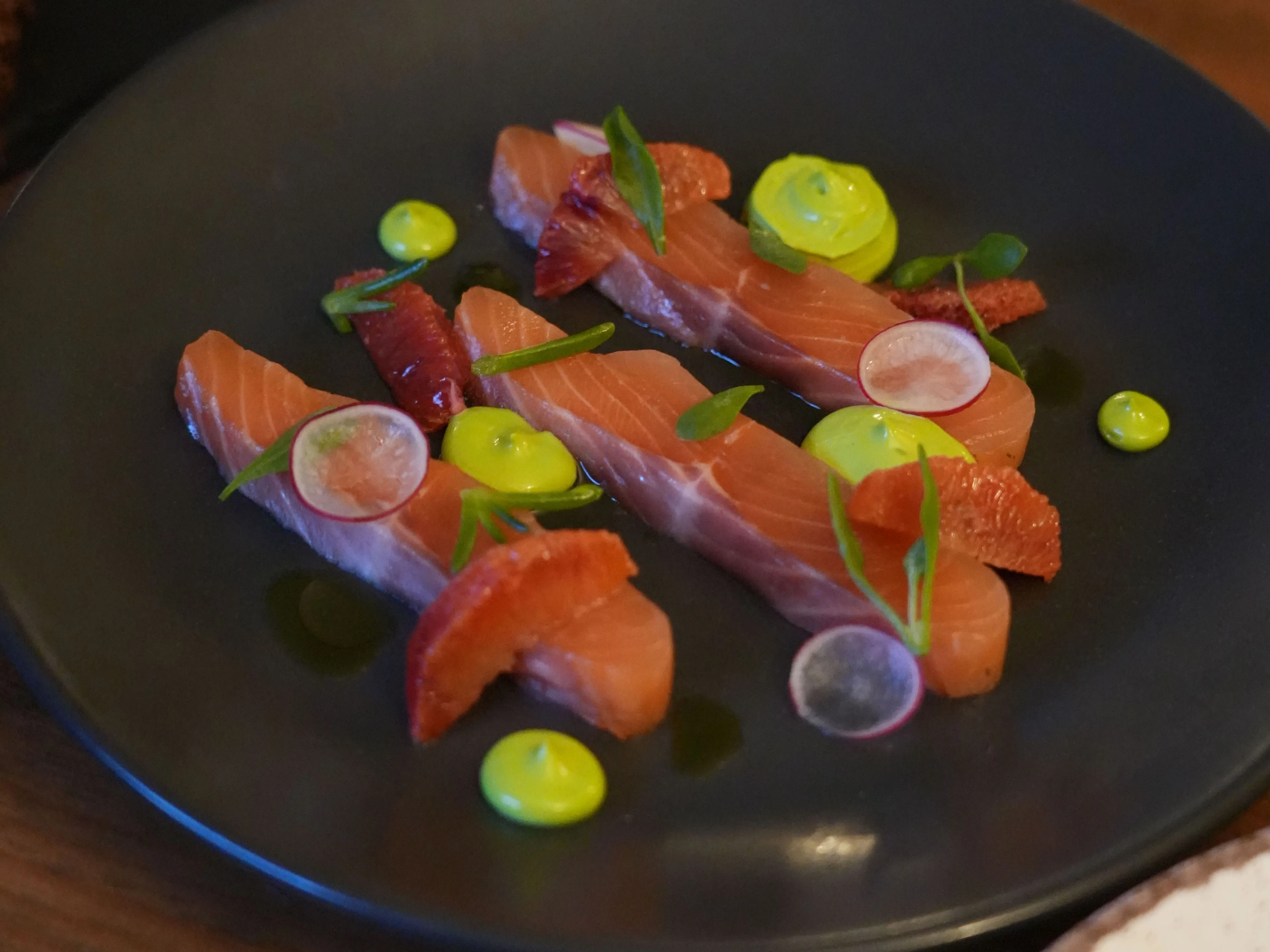 Artfully plated salmon sashimi with citrus, radish, herbs and bright green sauce on a black plate.
