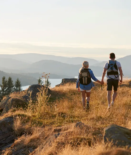Couple with backpacks hiking hand in hand along a sunlit hillside above a lake and misty mountains