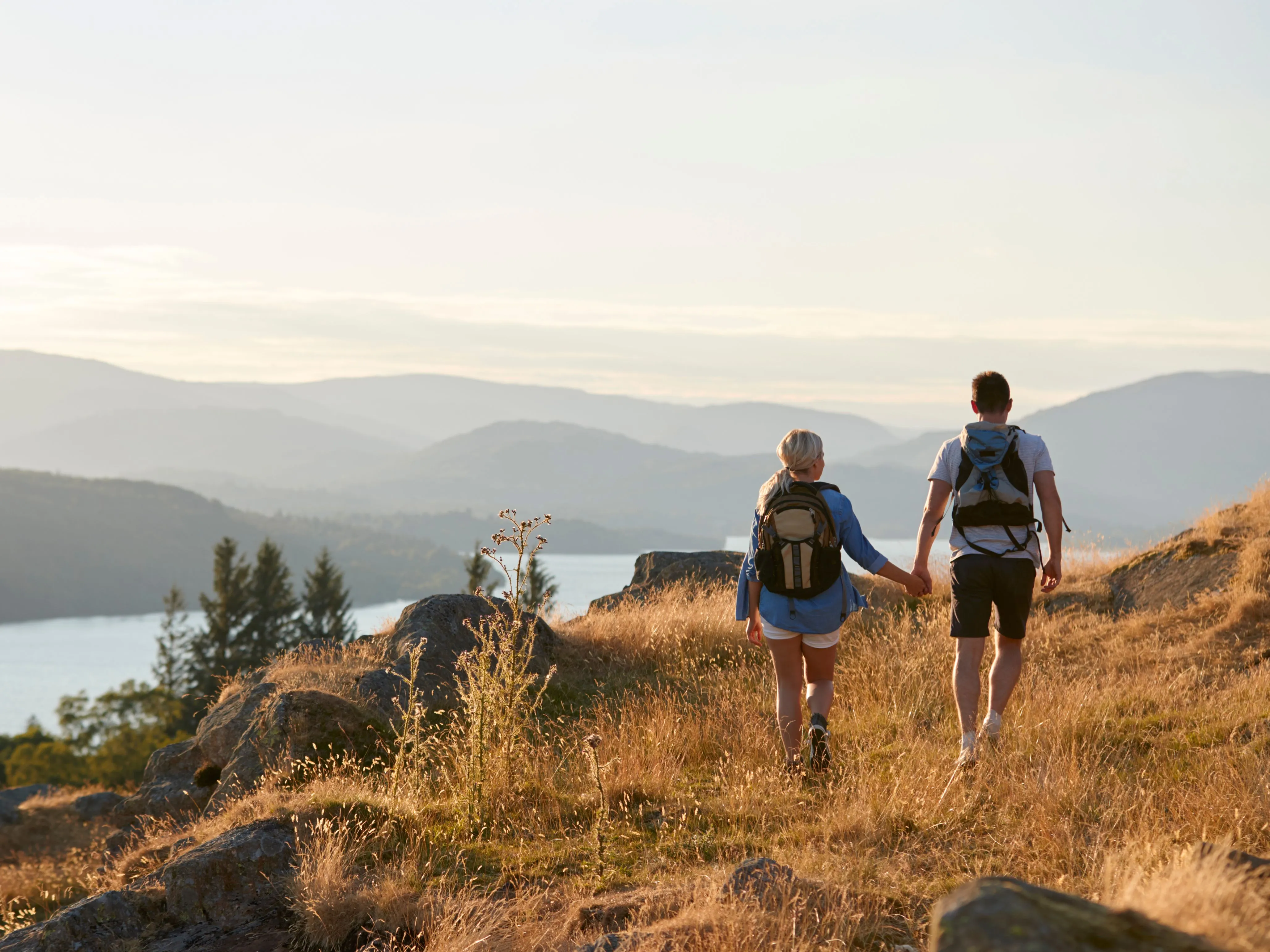Couple with backpacks hiking hand in hand along a sunlit hillside above a lake and misty mountains