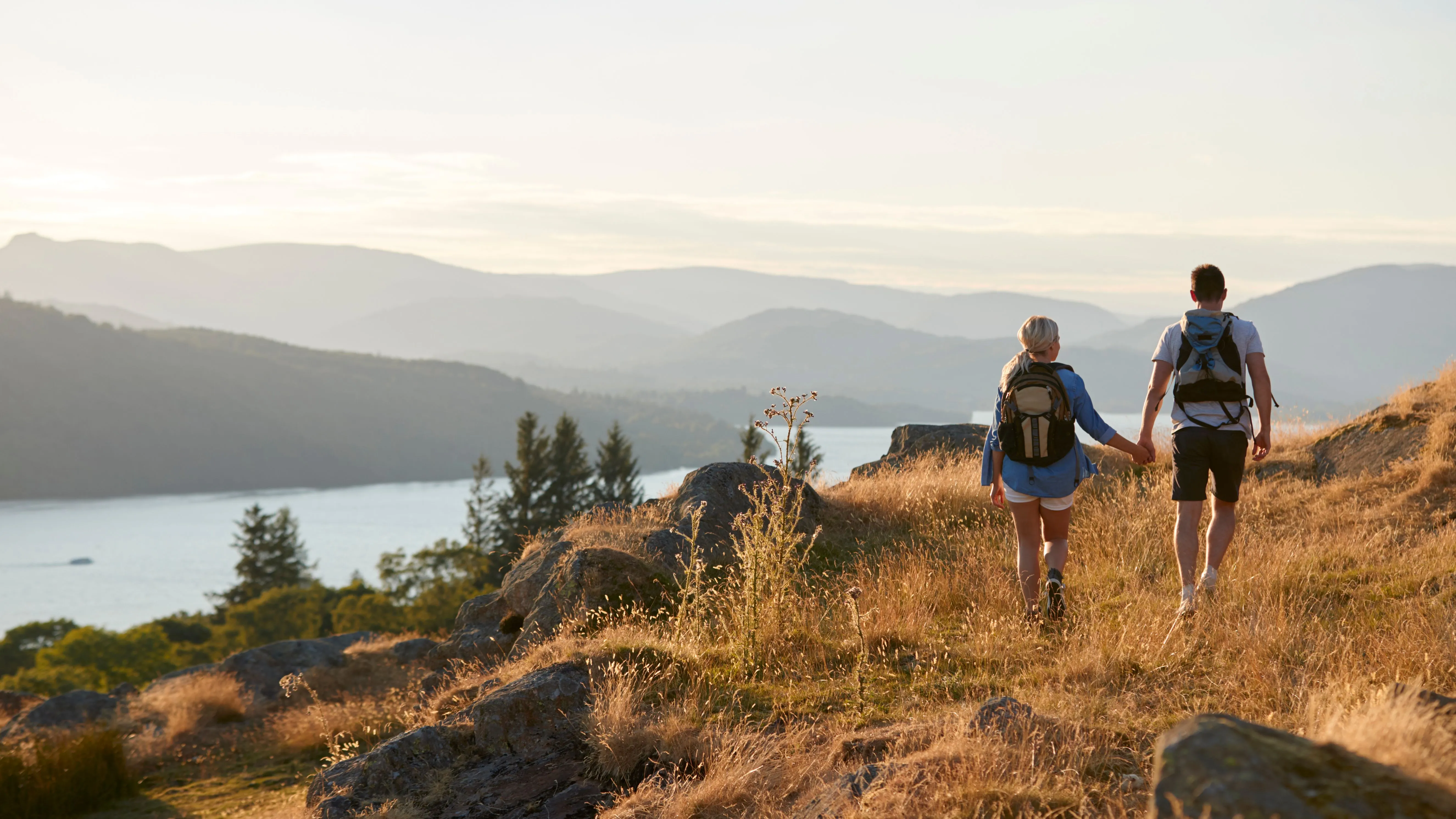 Couple with backpacks hiking hand in hand along a sunlit hillside above a lake and misty mountains