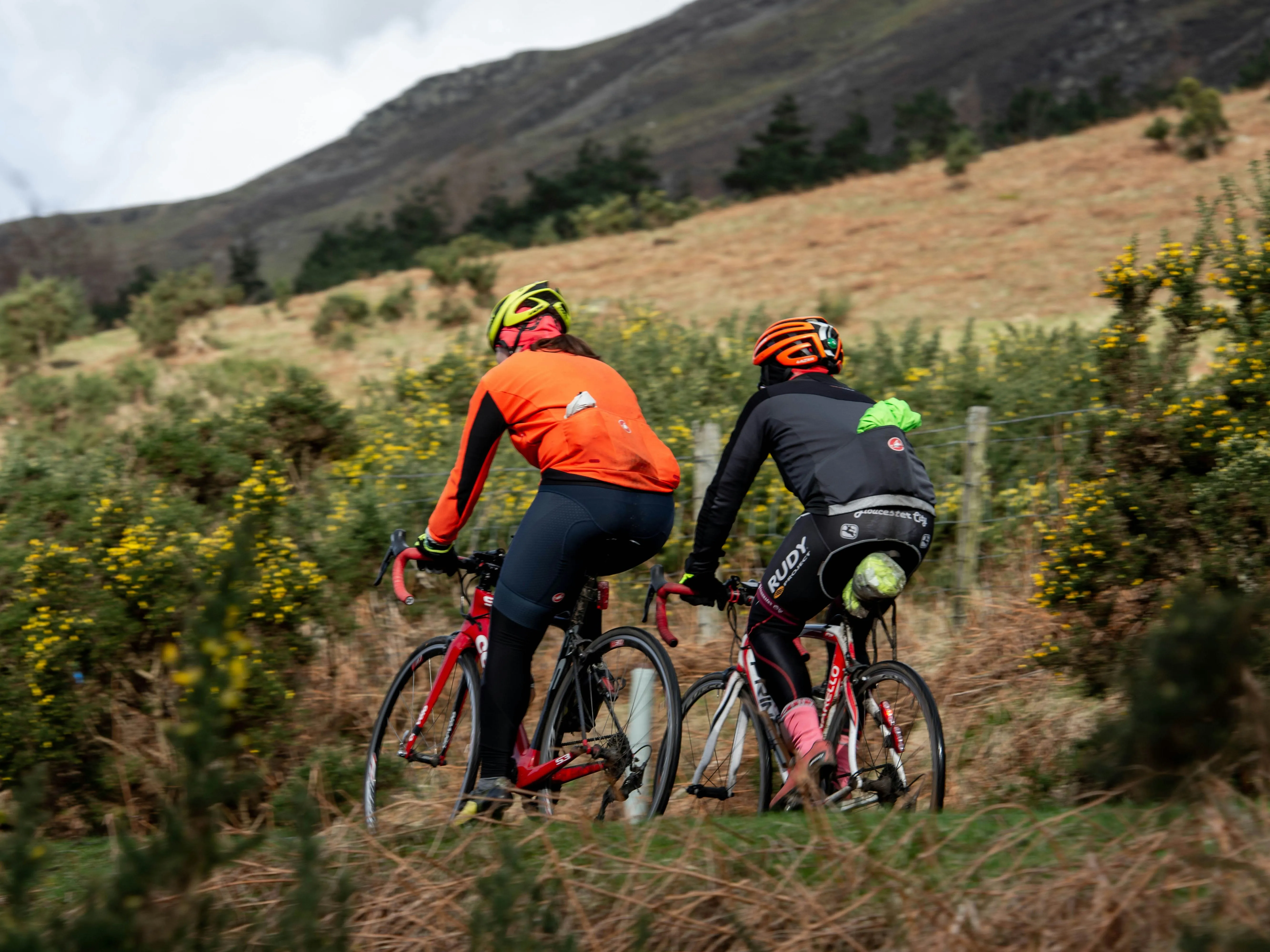 Two cyclists in bright helmets and jackets riding road bikes through a mountainous rural landscape.