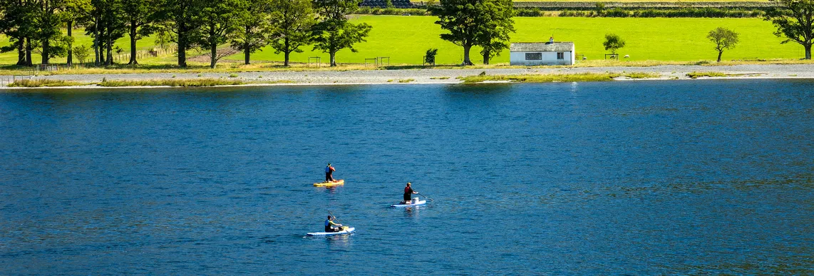 Three paddleboarders on a blue lake beneath bright green mountain slopes and a small white cottage.