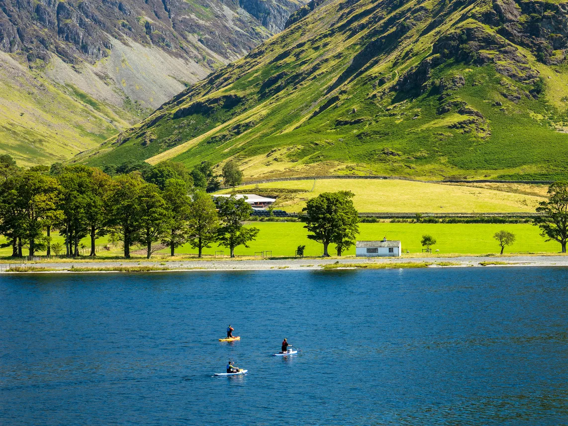 Three paddleboarders on a blue lake beneath bright green mountain slopes and a small white cottage.