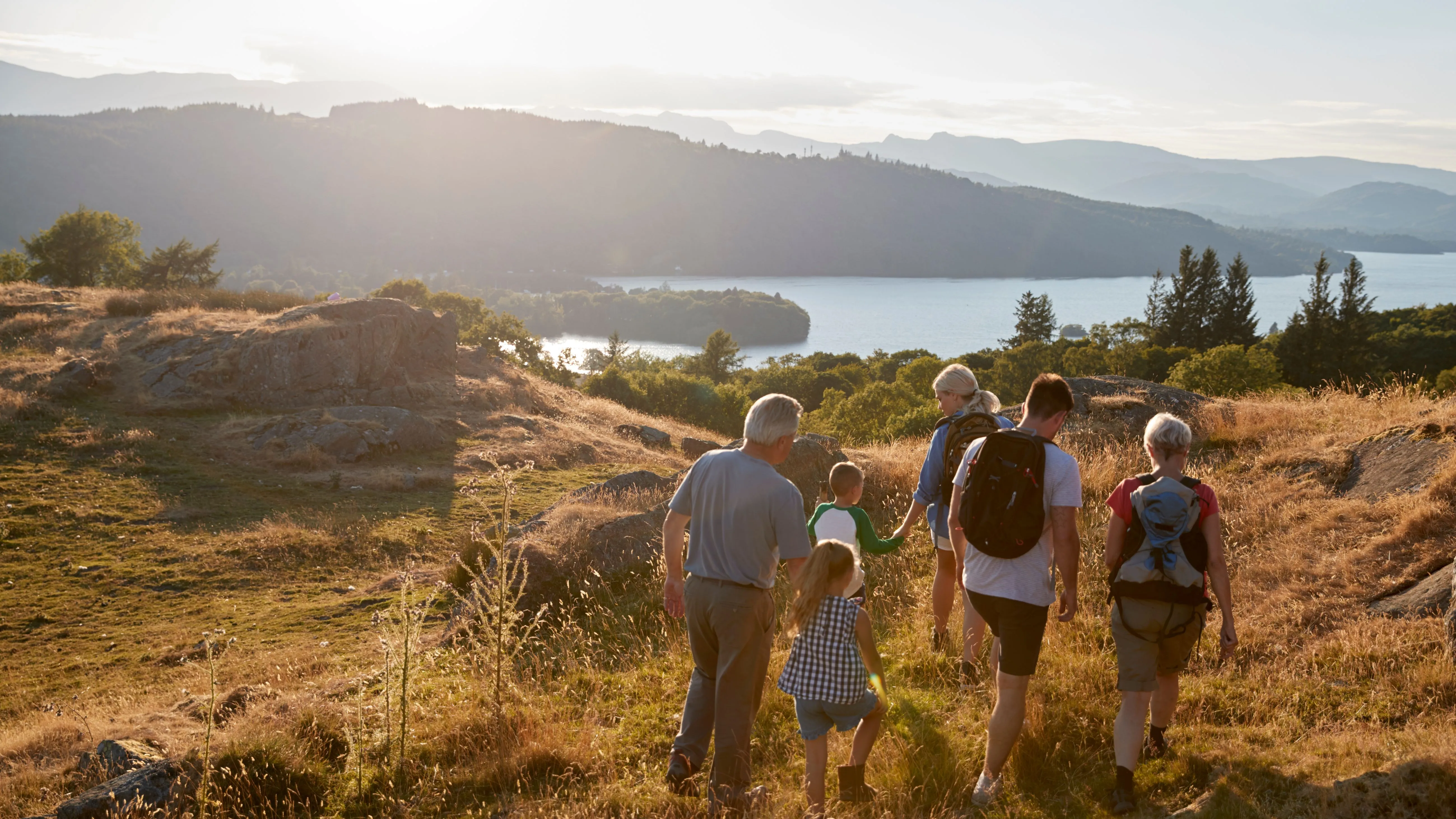 Multi-generational family hiking down a sunlit hillside overlooking a lake and layered mountains.