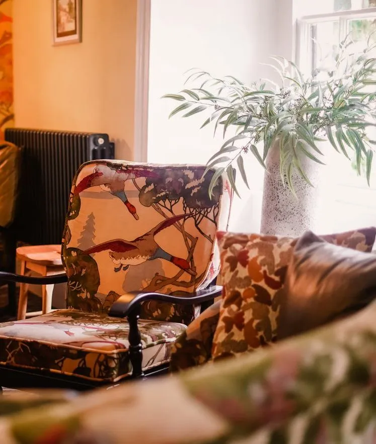 Cosy eclectic sitting room with patterned armchairs, floral wallpaper and a potted plant by the window.