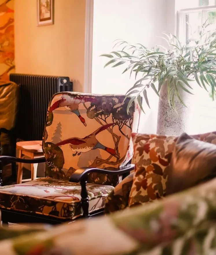 Cosy eclectic sitting room with patterned armchairs, floral wallpaper and a potted plant by the window.
