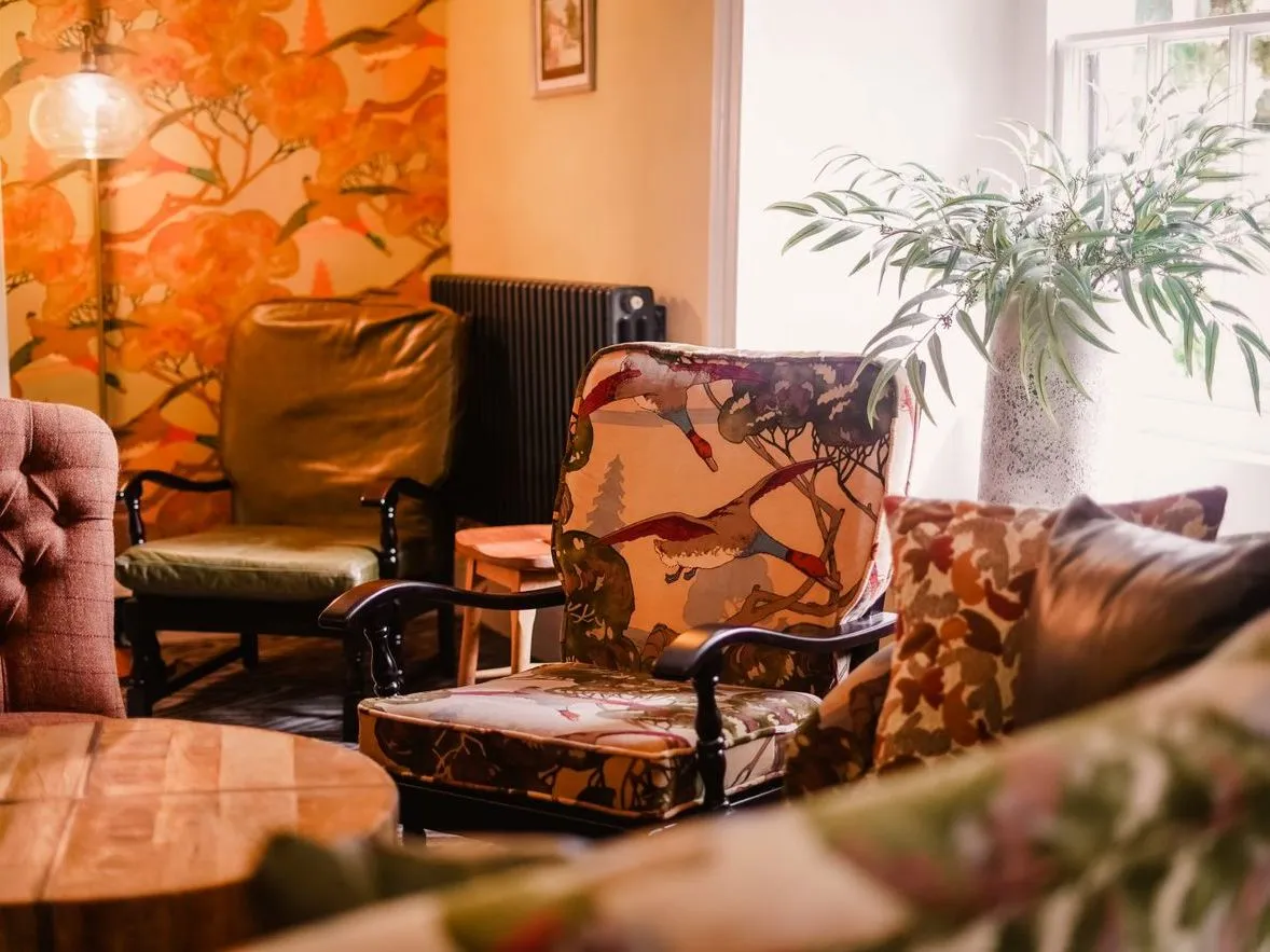 Cosy eclectic sitting room with patterned armchairs, floral wallpaper and a potted plant by the window.