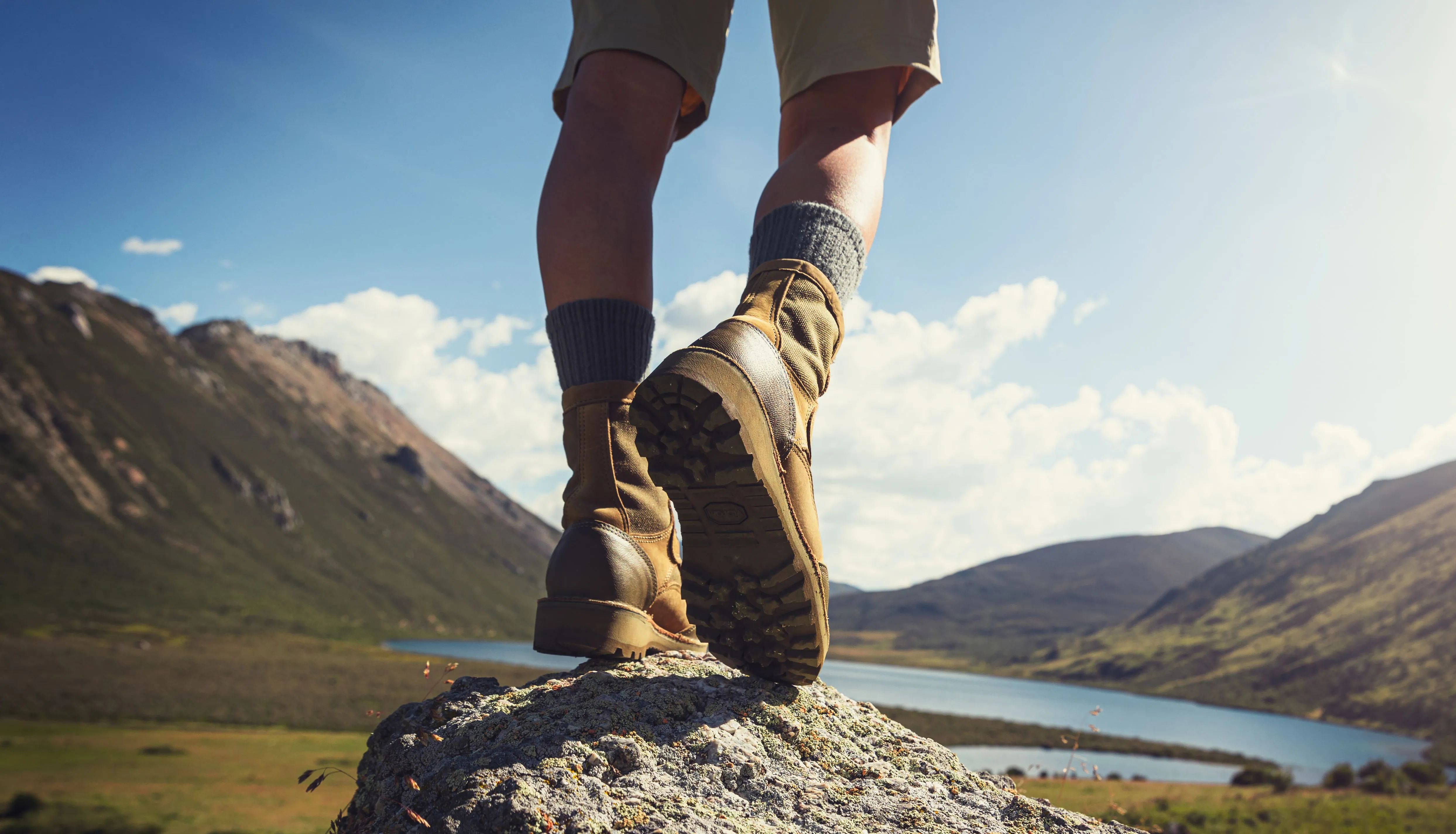 Low-angle view of a hiker in walking boots standing on a rock above a mountain lake.