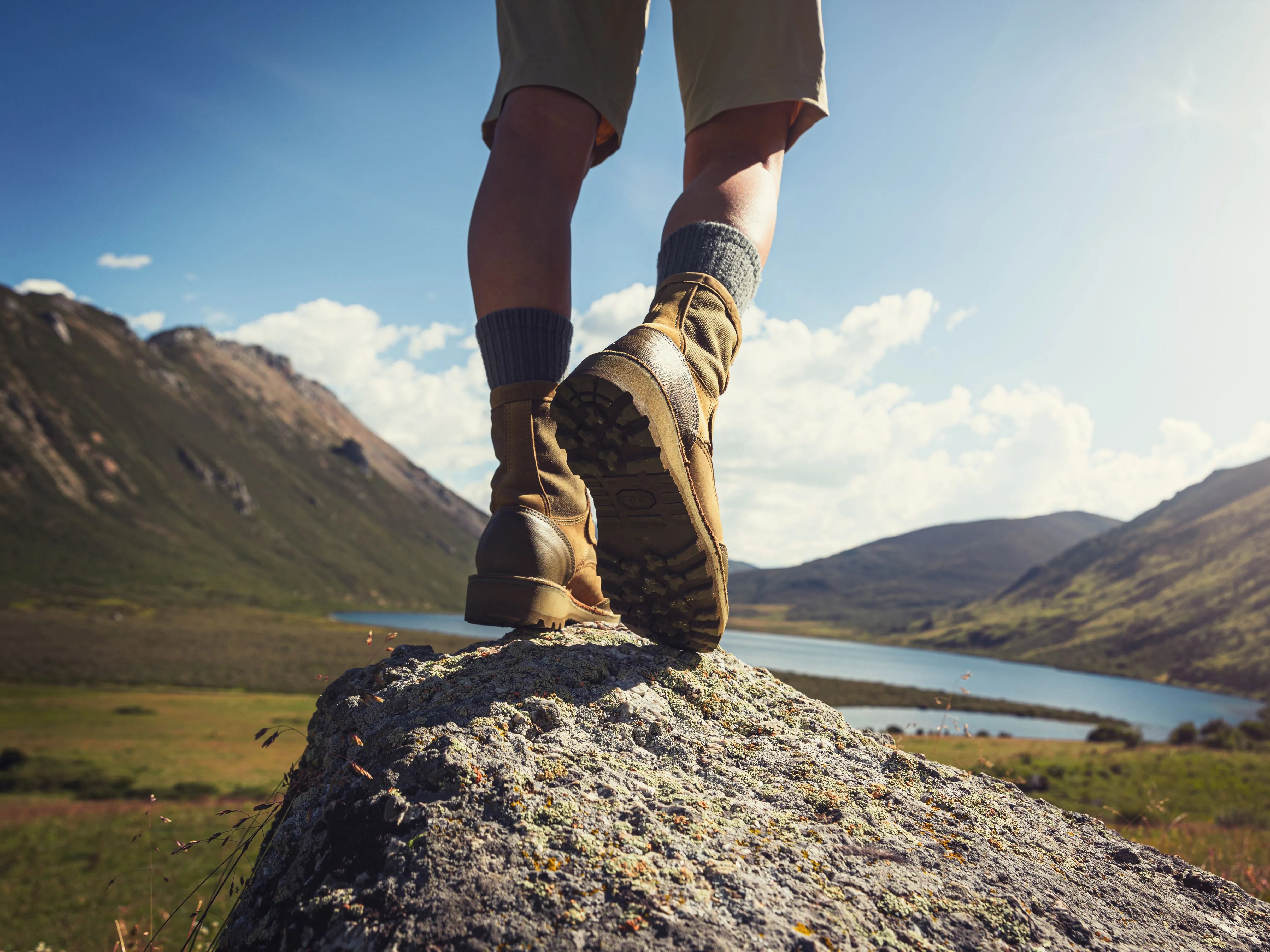 Low-angle view of a hiker in walking boots standing on a rock above a mountain lake.