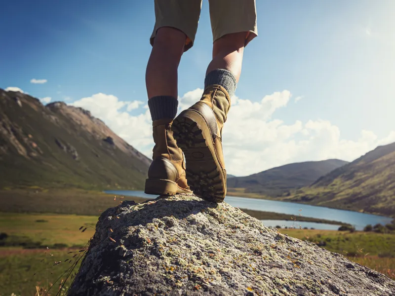 Low-angle view of a hiker in walking boots standing on a rock above a mountain lake.