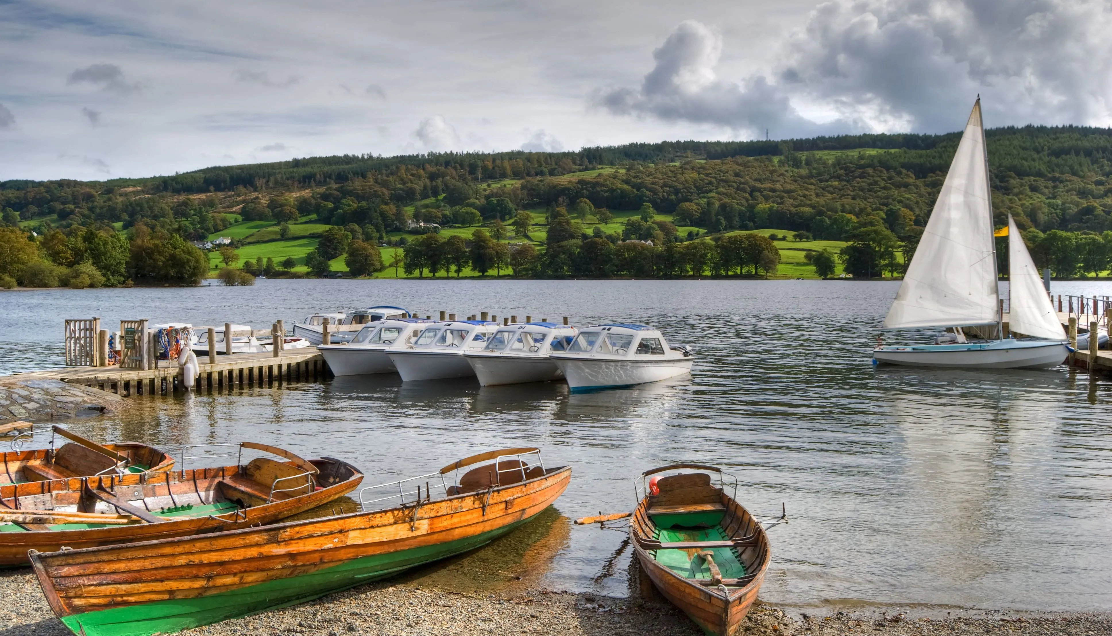 Wooden rowing boats, motorboats and sailing boats moored on a lake beneath green hills and dramatic clouds.