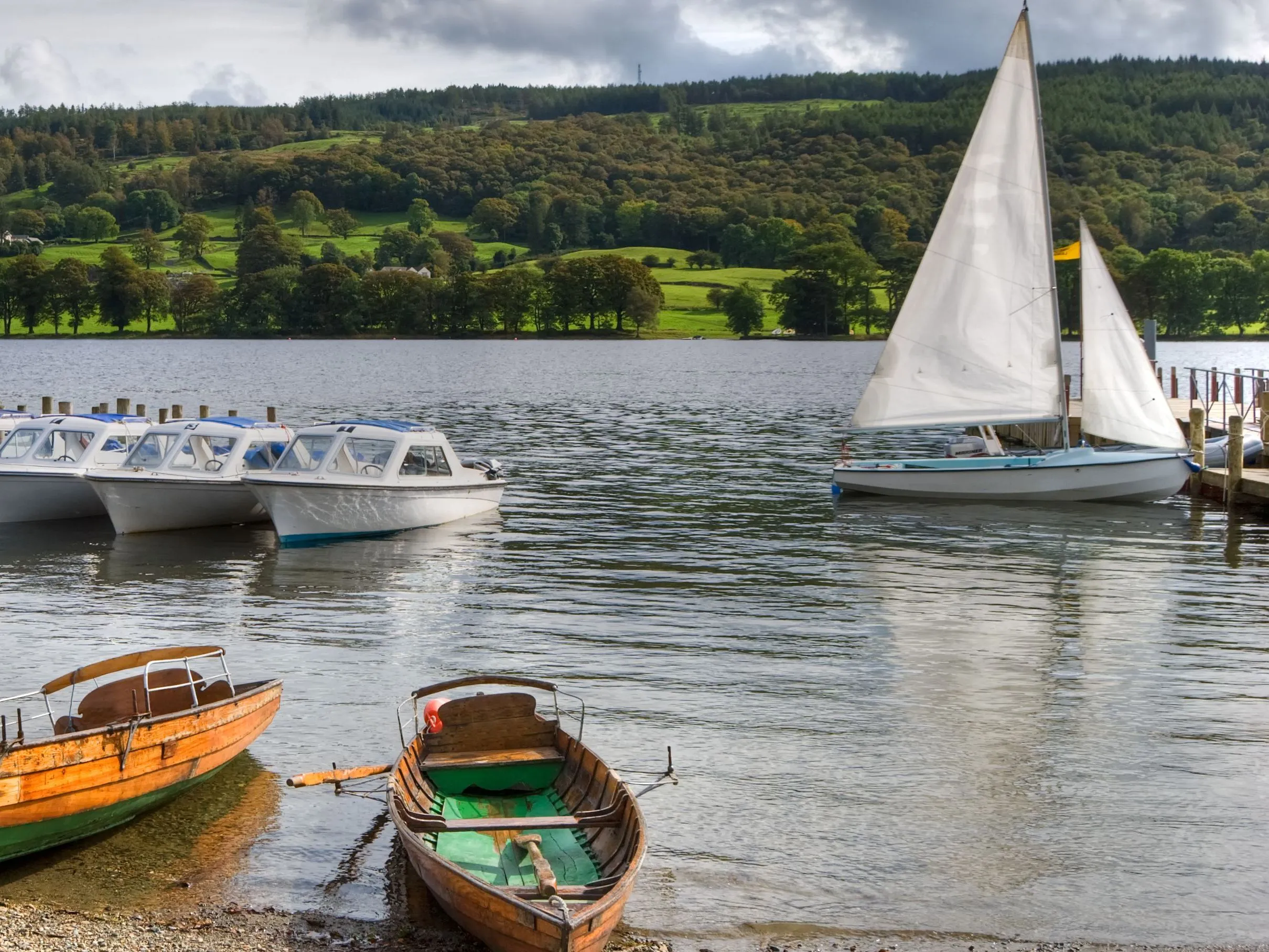 Wooden rowing boats, motorboats and sailing boats moored on a lake beneath green hills and dramatic clouds.