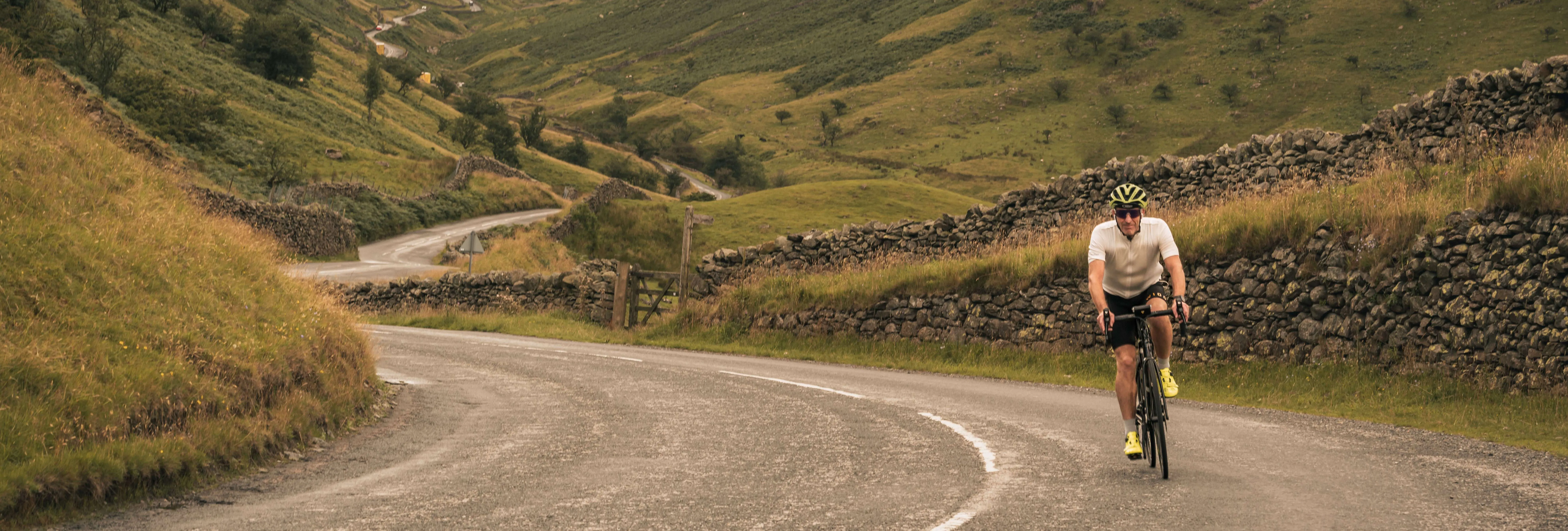 Cyclist riding along a winding country road through a green mountain valley bordered by dry-stone walls.