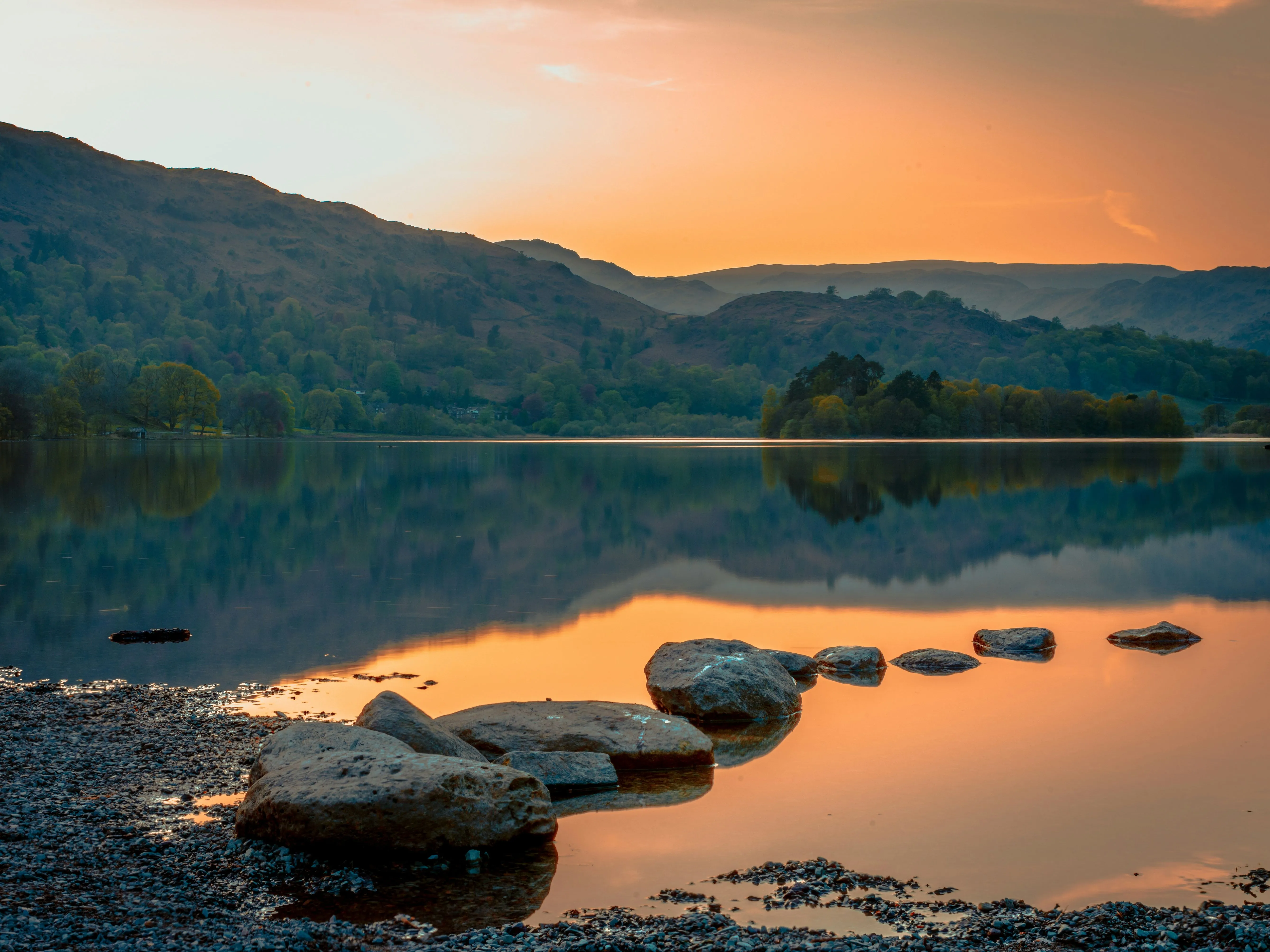 Sunset over a tranquil mountain lake with rocks in the foreground and hills reflected in still water