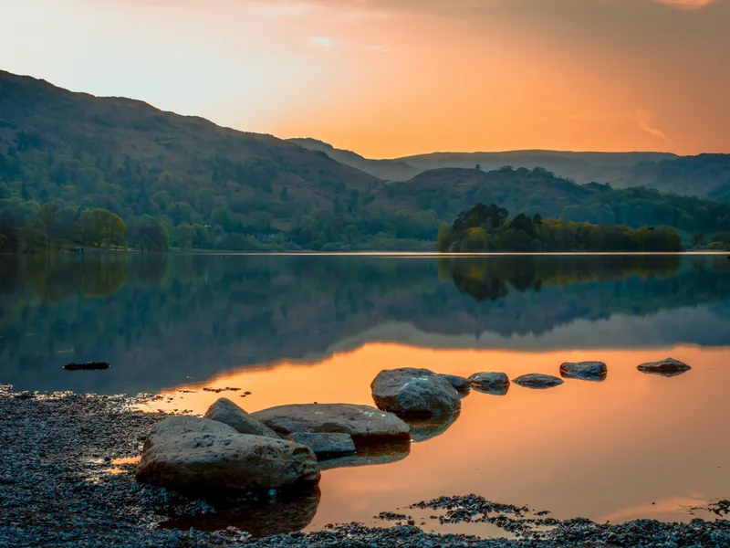 Sunset over a tranquil mountain lake with rocks in the foreground and hills reflected in still water