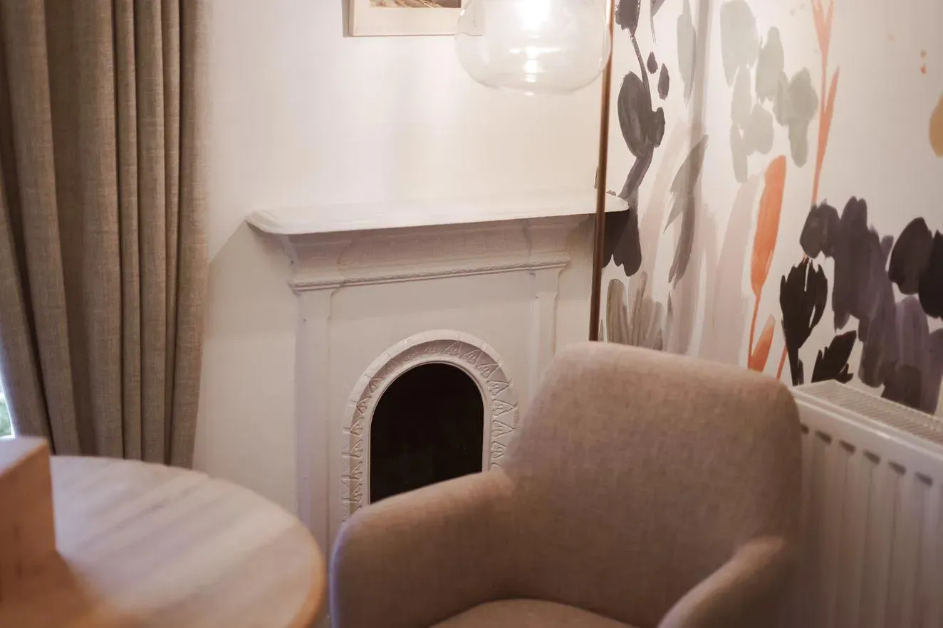 Cosy interior corner with a beige armchair, decorative fireplace, pendant light and floral wallpaper.