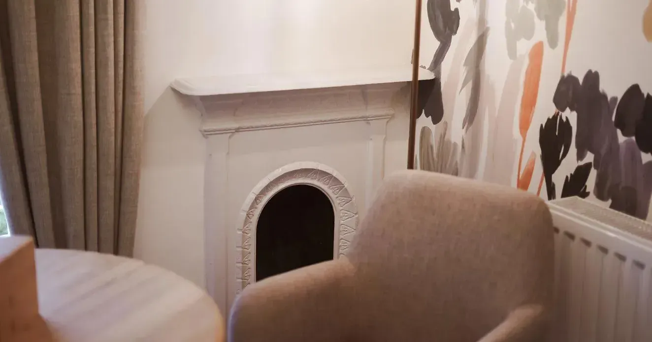 Cosy interior corner with a beige armchair, decorative fireplace, pendant light and floral wallpaper.