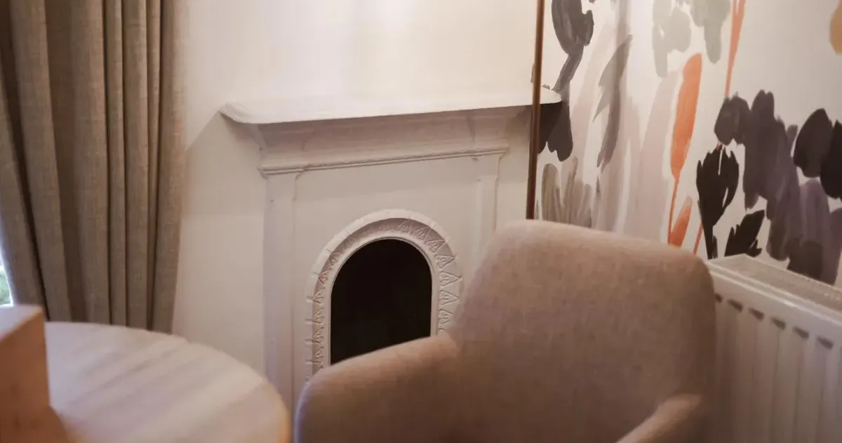 Cosy interior corner with a beige armchair, decorative fireplace, pendant light and floral wallpaper.