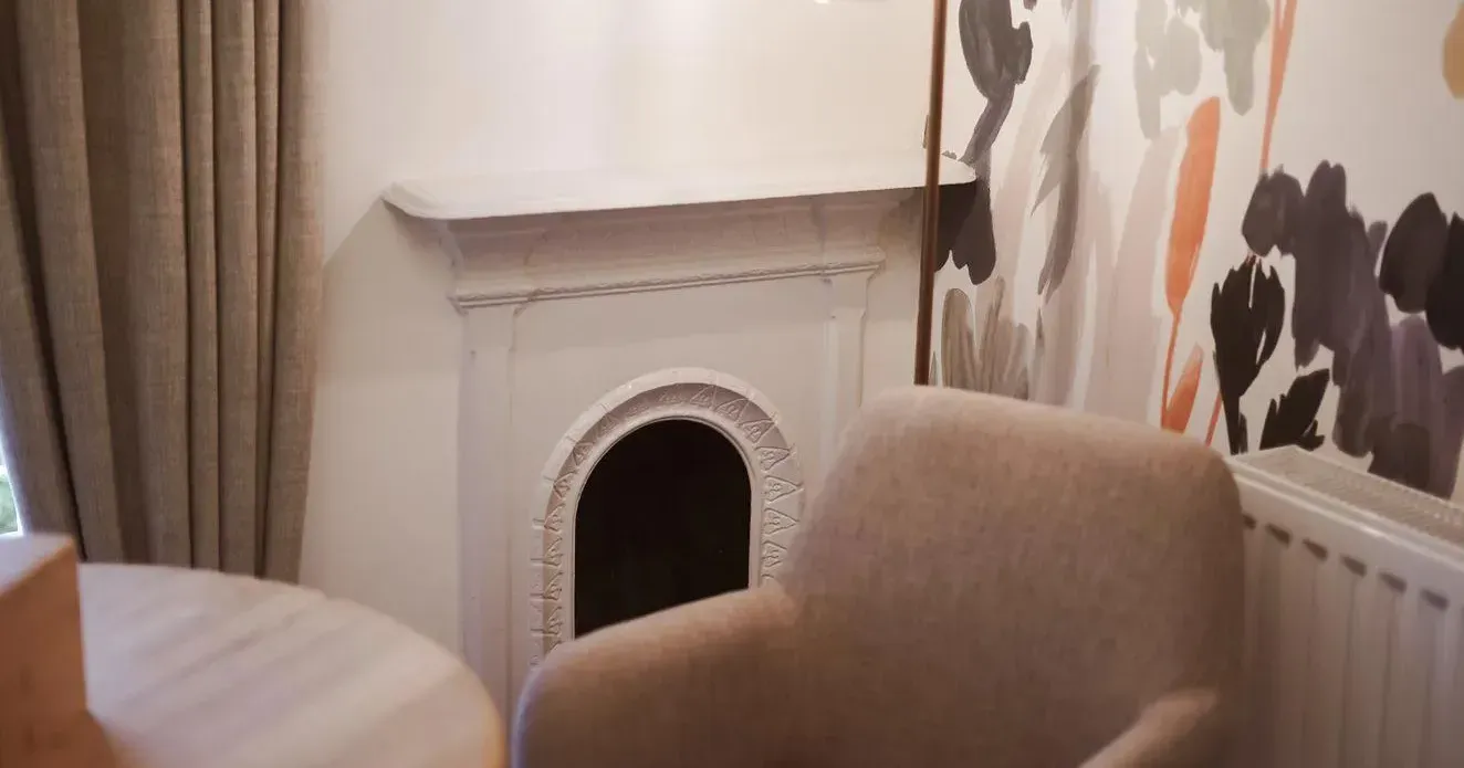 Cosy interior corner with a beige armchair, decorative fireplace, pendant light and floral wallpaper.
