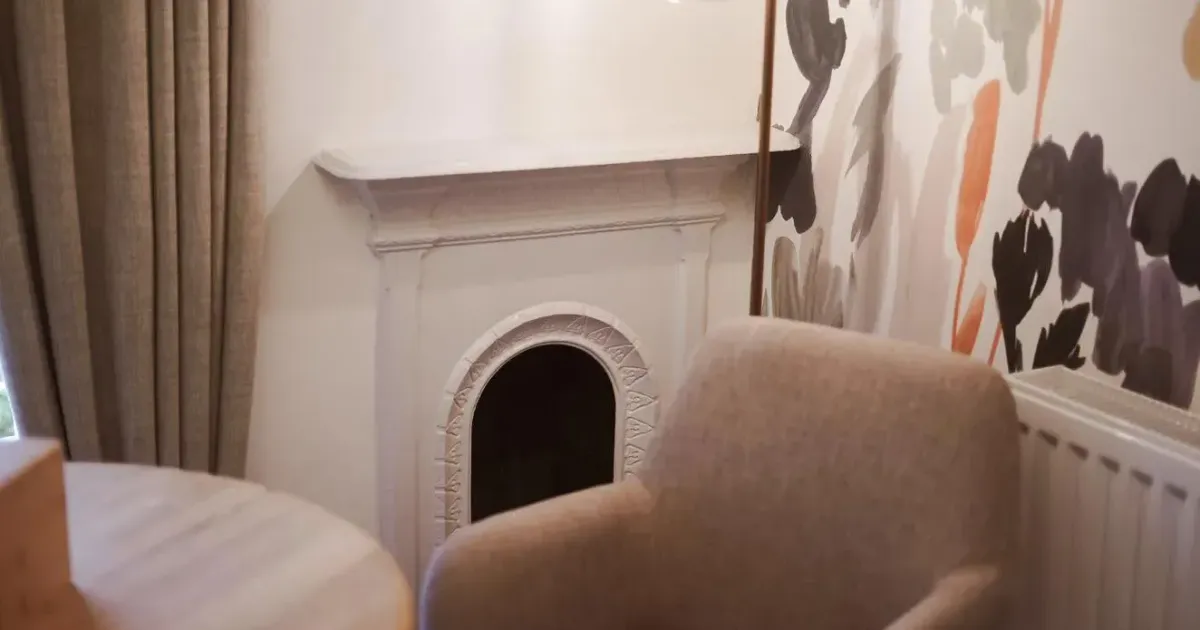 Cosy interior corner with a beige armchair, decorative fireplace, pendant light and floral wallpaper.