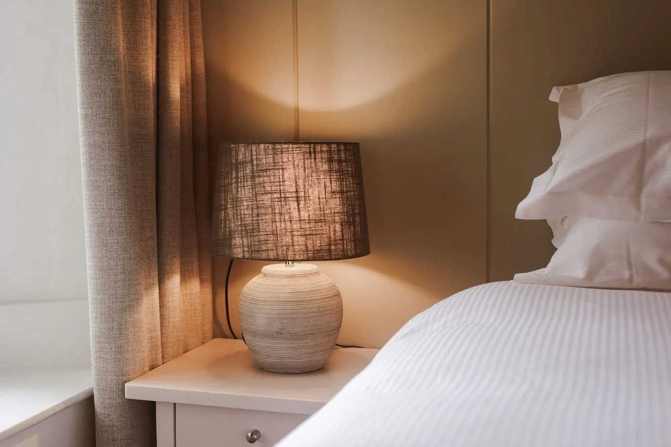 Warm bedside lamp on a white bedside table beside a neatly made bed and curtains.