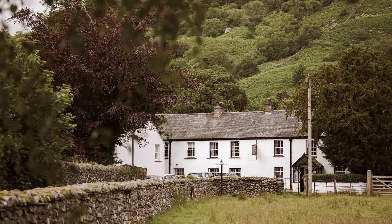 White country inn beneath a lush green hillside, framed by trees and dry-stone walls.