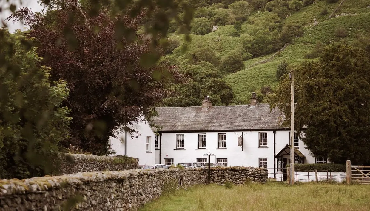 White country inn beneath a lush green hillside, framed by trees and dry-stone walls.