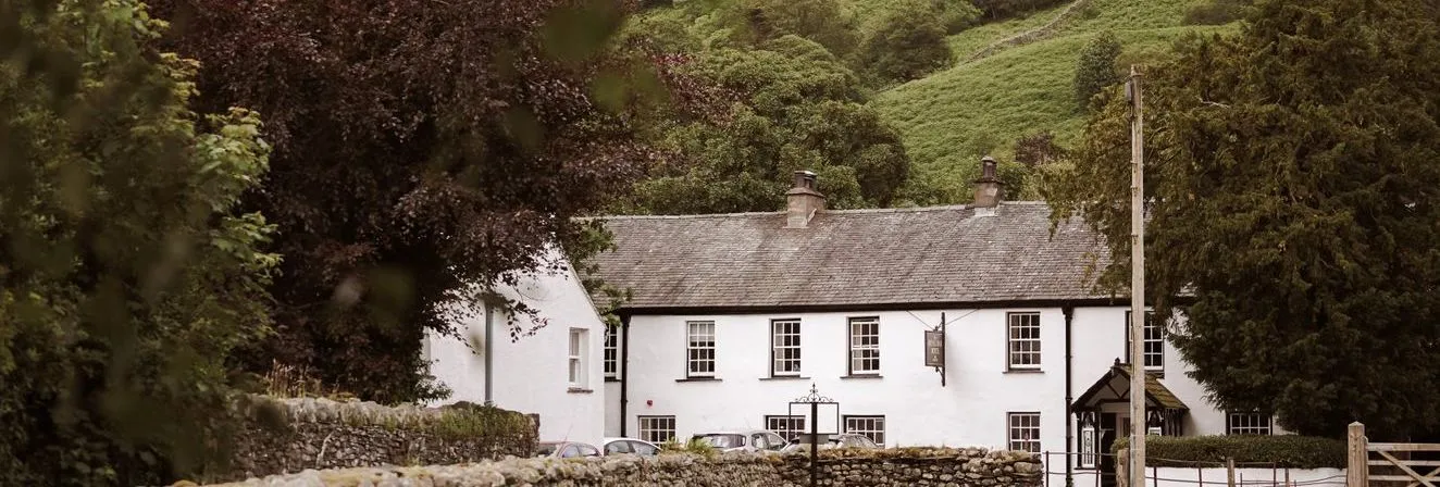 White country inn beneath a lush green hillside, framed by trees and dry-stone walls.