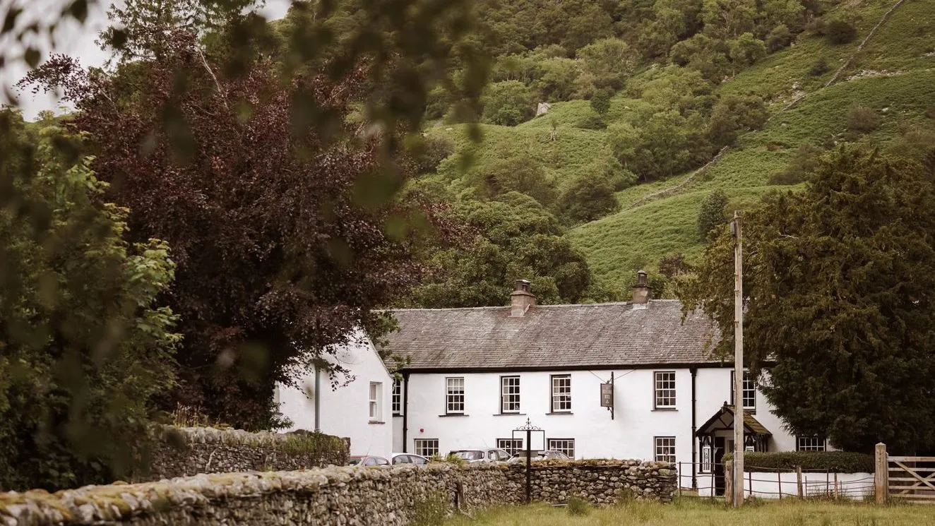 White country inn beneath a lush green hillside, framed by trees and dry-stone walls.