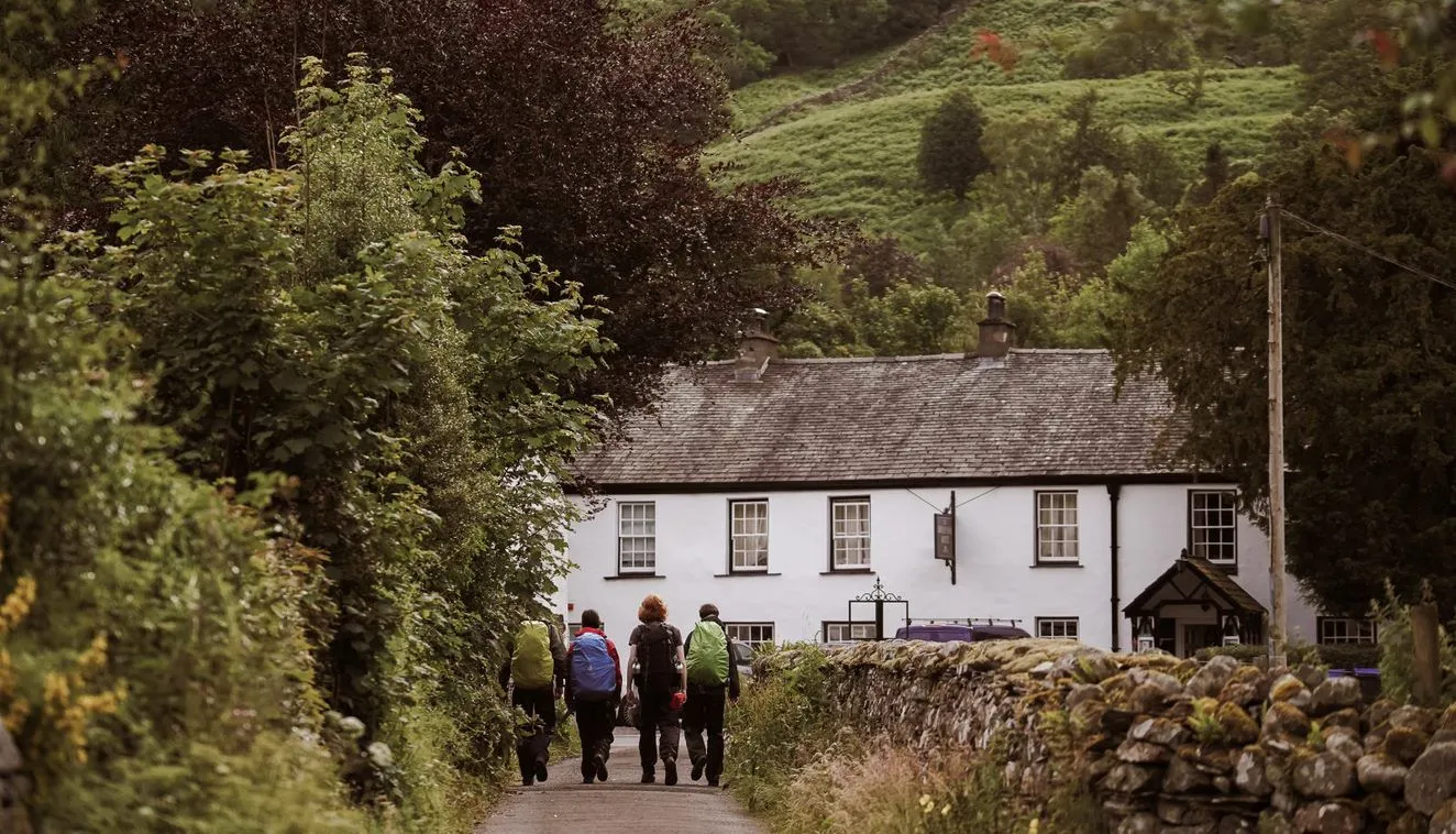 Three hikers with backpacks walking along a country lane towards a white cottage beneath lush green hills.