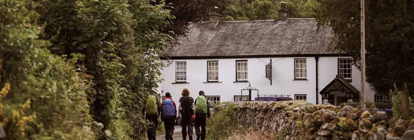 Three hikers with backpacks walking along a country lane towards a white cottage beneath lush green hills.