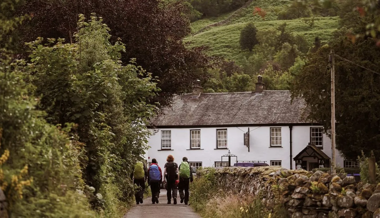 Three hikers with backpacks walking along a country lane towards a white cottage beneath lush green hills.