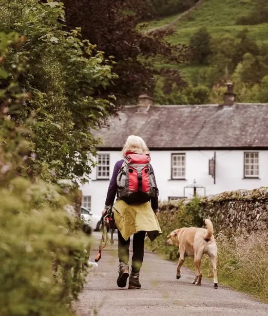 Hiker with a red backpack walking a dog along a leafy lane towards a white cottage.