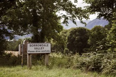 Roadside sign welcoming drivers to Borrowdale Valley amid leafy trees and distant hills.