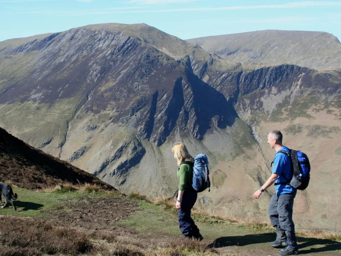 Two hikers with backpacks and a dog walking along a mountain ridge above a rugged valley.