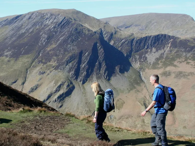 Two hikers with backpacks and a dog walking along a mountain ridge above a rugged valley.