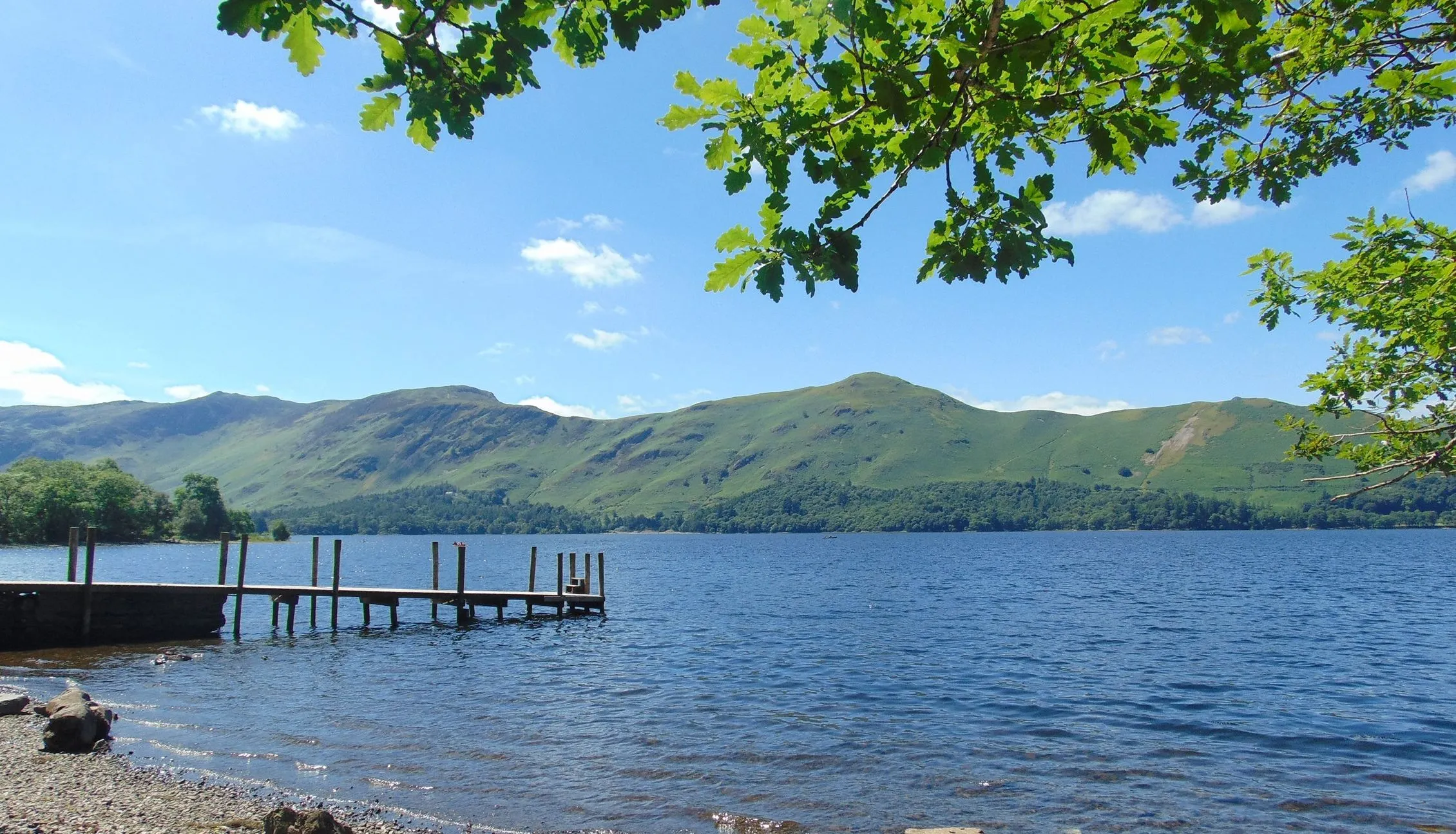 Wooden jetty beside a clear blue lake with green hills beneath leafy tree branches.
