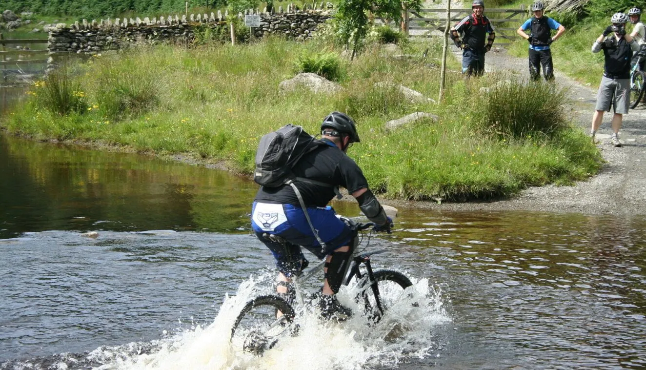 Mountain biker splashing through a shallow river while fellow cyclists watch from a rural track